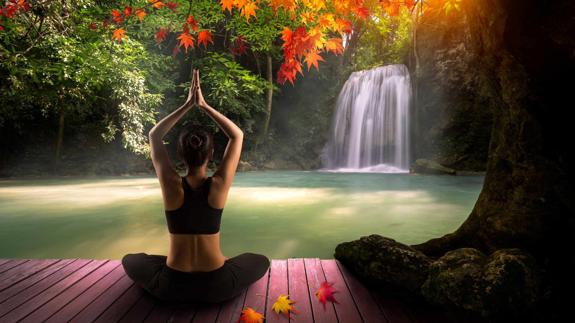 A white woman lifting her arms in a yoga pose, while sitting on a pink dock, in front of a small waterfall and pond.